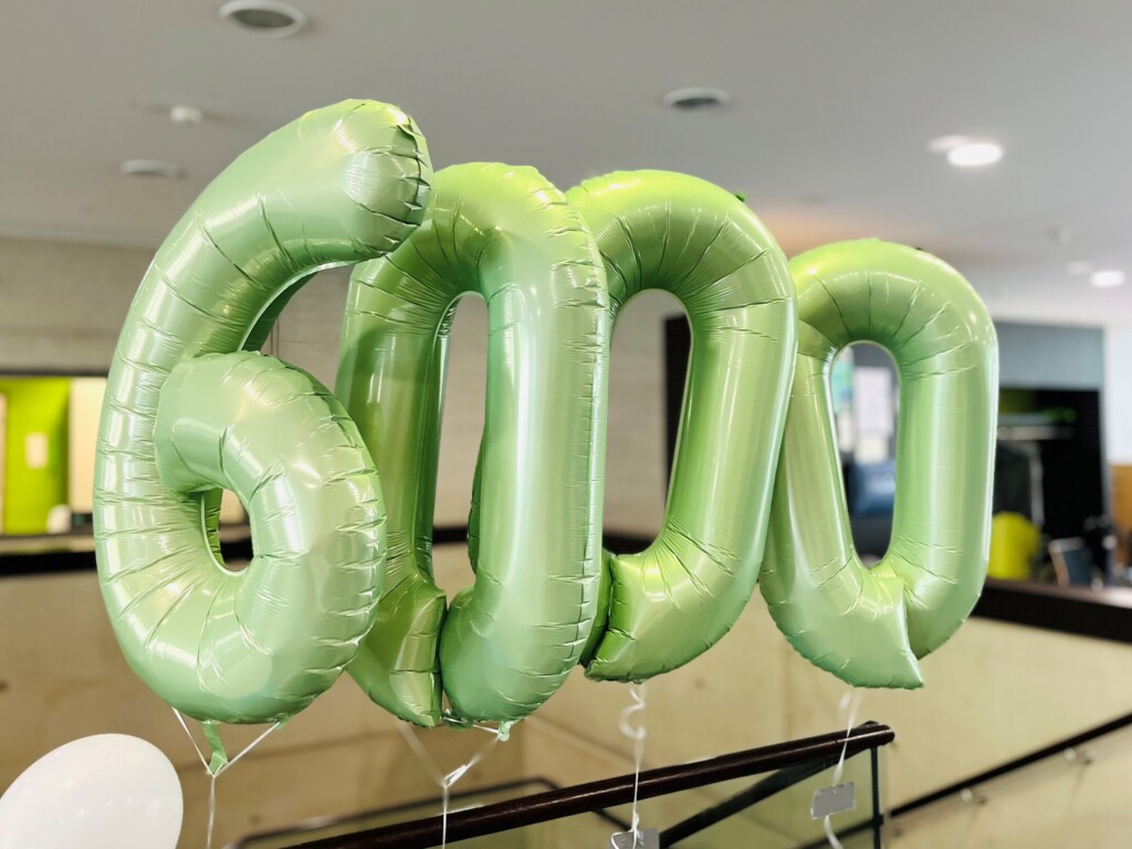 Nahaufnahme grüner Folienluftballons in Form der Zahl 6000 vor einem Innenraum mit heller Decke.